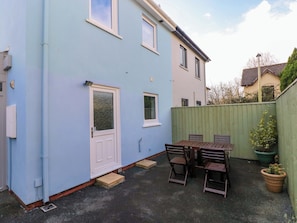 A two-story house with a blue exterior, a front door, a small patio with a table and chairs, and a green fence. - No.22 The Clicketts (Tenby)