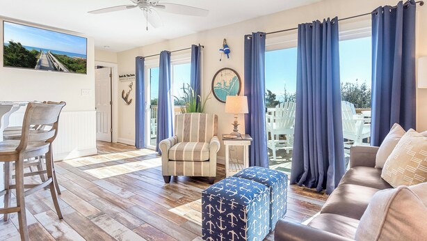 Una habitación con ventilador de techo, zona de comedor con sillas, banco acolchado, sofá y vistas amplias a través de ventanas con cortinas.