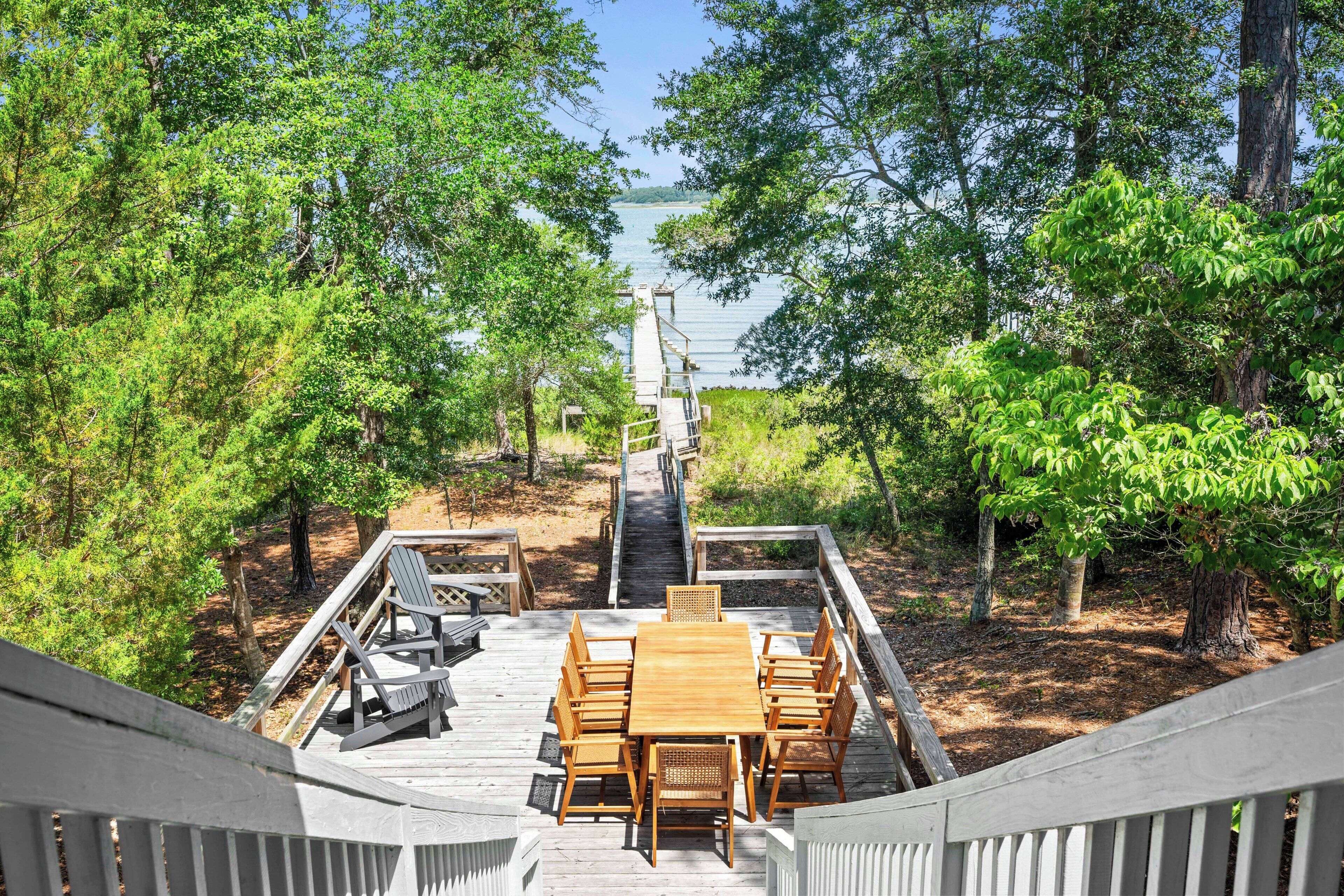 A wooden deck with a table and chairs, surrounded by trees, leading to a dock over water.