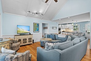A spacious living room with a large gray sofa, a television, and a dining area with wooden cabinets and a kitchen in the background. - Hark the Sound | Soundfront 4BR w/ Private Pier, Golf Cart & Sunset Views (Emerald Isle)