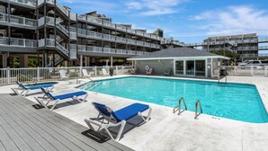 Ein Schwimmbecken mit Liegestühlen, einem Rettungsschwimmerstand und einem Gebäude mit mehreren Balkonen.
