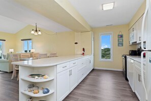 A kitchen with white cabinets, a central island, and a dining area with a table and chairs. - Colony by the Sea 301 | Oceanview 2BR Condo w/ Pool, Hot Tub & Beach Access (Indian Beach)