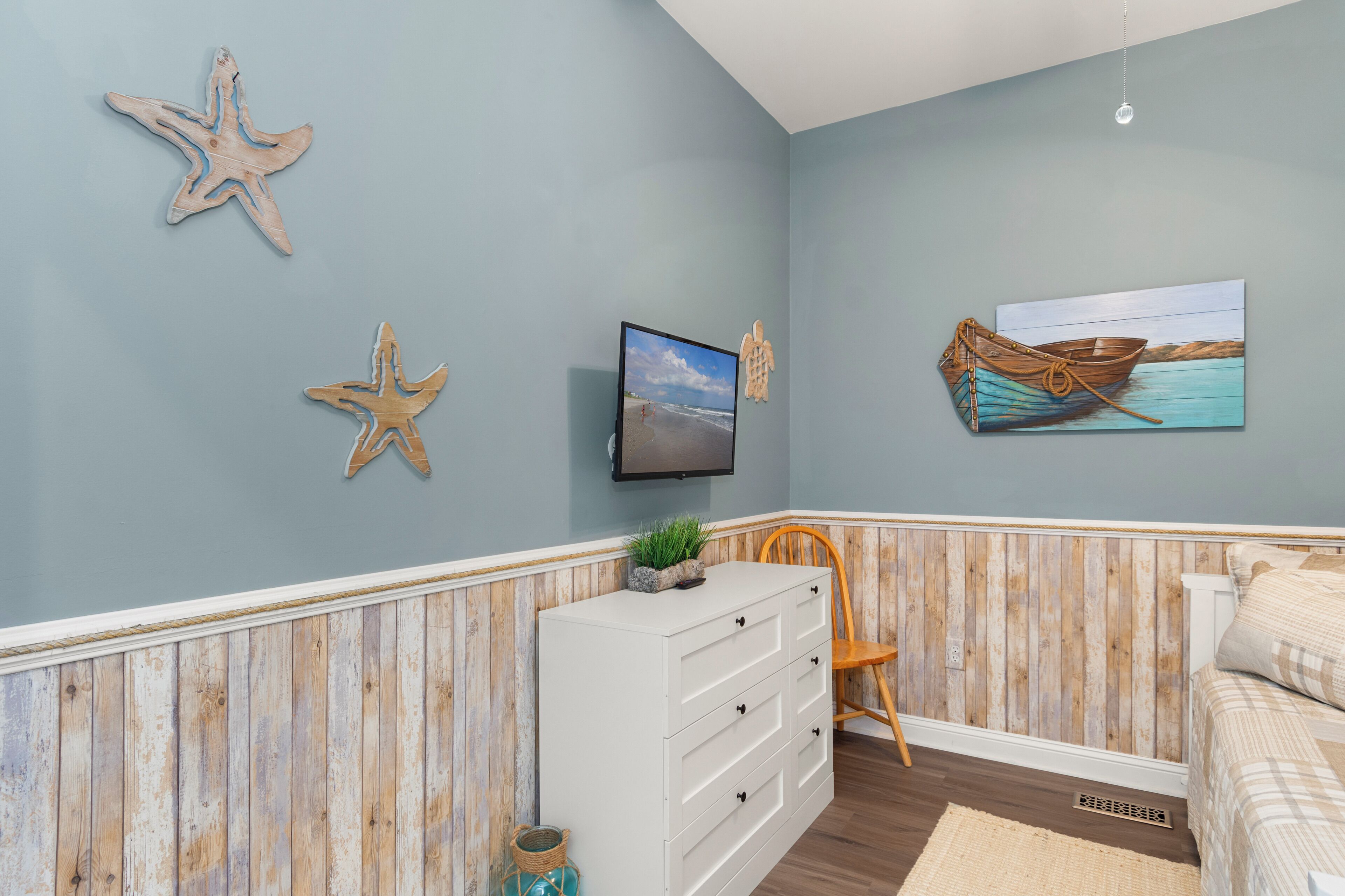 A bedroom with a wooden wall panel, a white dresser, a television, and a painting of a boat on water.