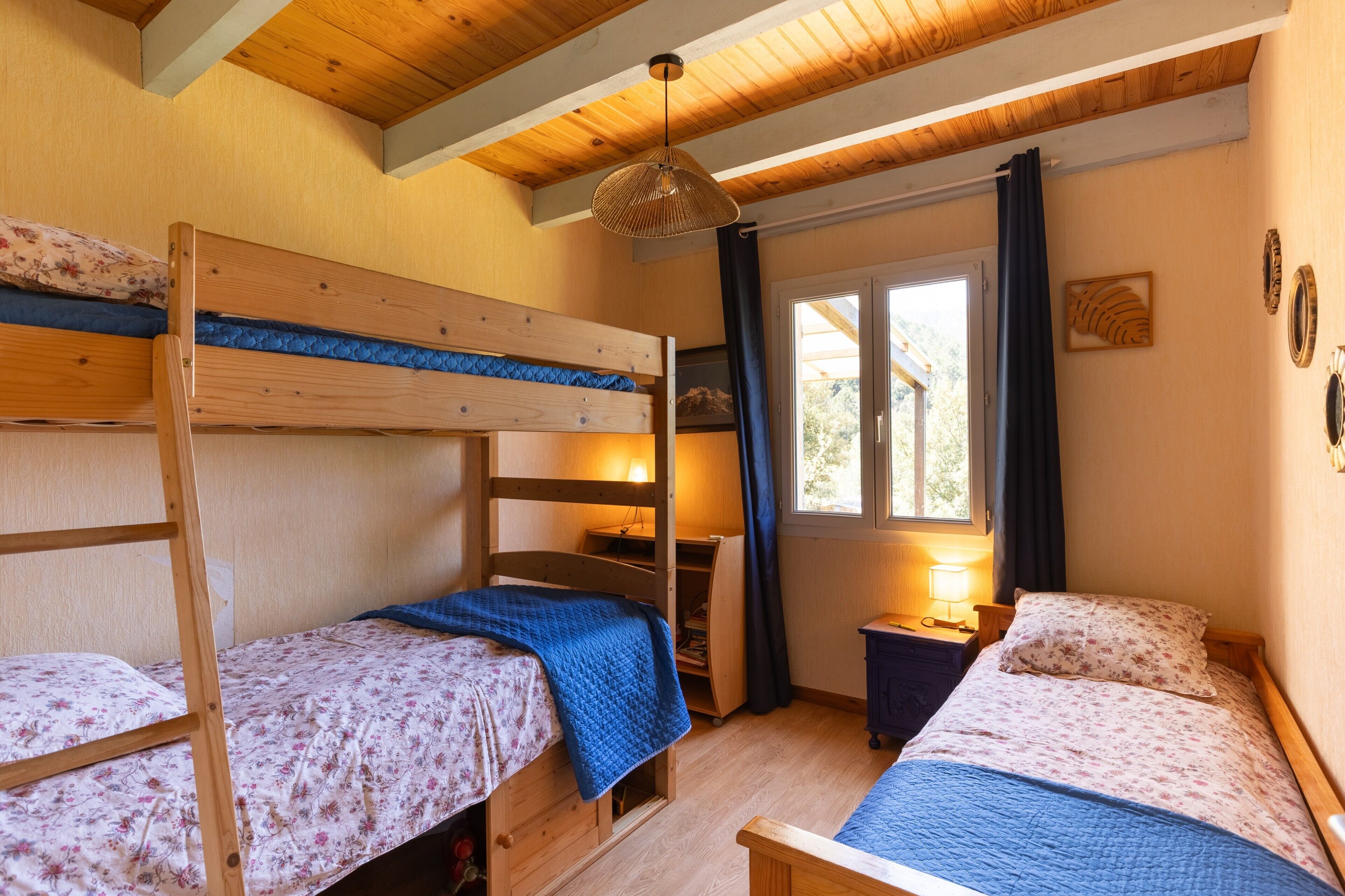 Ein Zimmer mit zwei Etagenbetten, einem Schreibtisch, einem Stuhl, einem Fenster mit Vorhängen und einer Pendelleuchte.