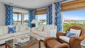 Quarto com dois sofás brancos, uma cadeira de vime, uma mesa de centro de vidro e grandes janelas com cortinas azuis.