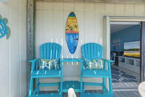 A room with two turquoise chairs, a surfboard art piece, and a view of the ocean. - Queens Court 1102 | Oceanfront 1BR Condo w/ Pool, Balcony & Beach Access (Emerald Isle)