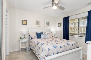 Un dormitorio con cama, mesitas de noche, ventilador de techo y ventana con cortinas.