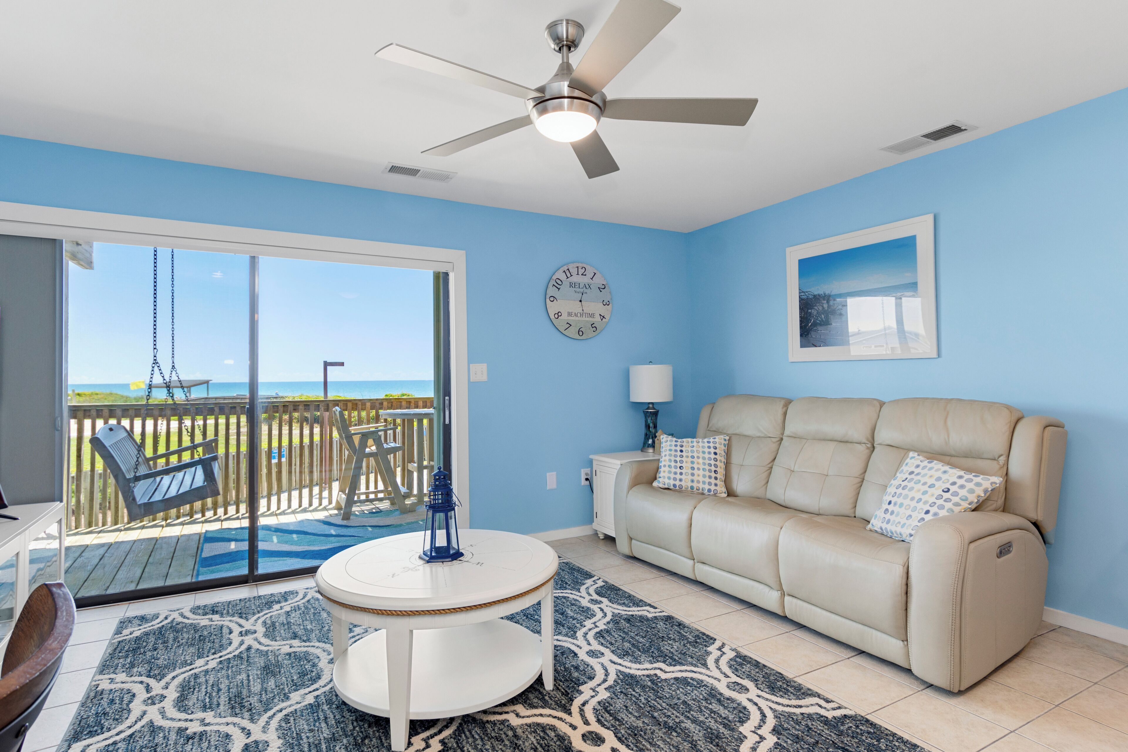 A room with a ceiling fan, a sofa, a table, a clock, and a view through a sliding glass door to an outdoor deck.