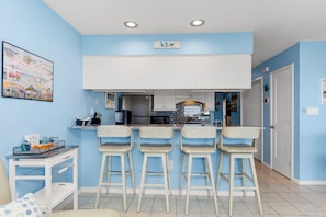 A kitchen with a blue wall, white cabinets, and three wicker bar stools. - Queens Court 1102 | Oceanfront 1BR Condo w/ Pool, Balcony & Beach Access (Emerald Isle)