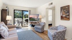 A living room with a sofa, armchairs, a TV, and a balcony with outdoor furniture. - Point Emerald Villa C-106 | Oceanview 2BR Condo w/ Pools, Tennis & Beach Access (Emerald Isle)