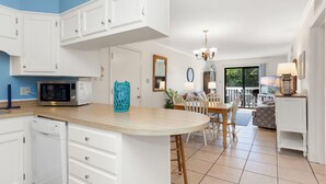 A kitchen with white cabinets, a microwave, and a dishwasher. There is a dining area with a table and chairs. - Point Emerald Villa C-106 | Oceanview 2BR Condo w/ Pools, Tennis & Beach Access (Emerald Isle)