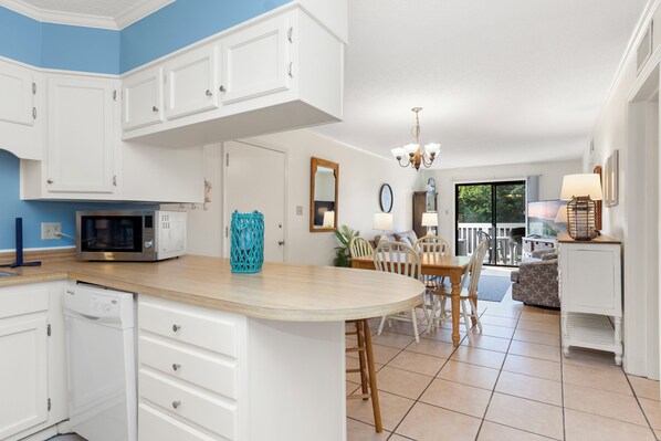 A kitchen with white cabinets, a microwave, and a dishwasher. There is a dining area with a table and chairs. - Point Emerald Villa C-106 | Oceanview 2BR Condo w/ Pools, Tennis & Beach Access (Emerald Isle)