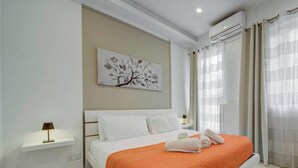 A hotel room with a bed, a nightstand, a wall-mounted air conditioner, and a window with curtains. - Msida 2BR Home Near University w/ Terrace & WiFi (Msida)