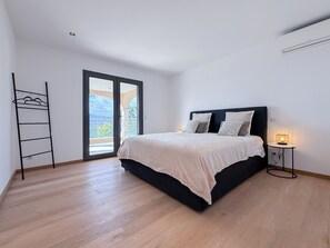 Une chambre à coucher comprenant un lit, une table de chevet, une lampe, une porte-fenêtre donnant sur un balcon et un étagère en métal.