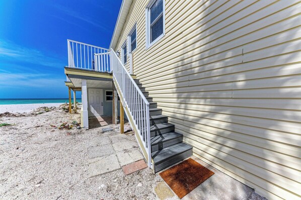 A beachfront house with a staircase leading to a balcony, a sandy beach, and a clear blue sky.