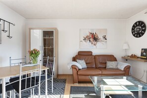 A living room with a brown leather sofa, a glass coffee table, a dining table with chairs, a vase with flowers, a painting on the wall, and a clock.