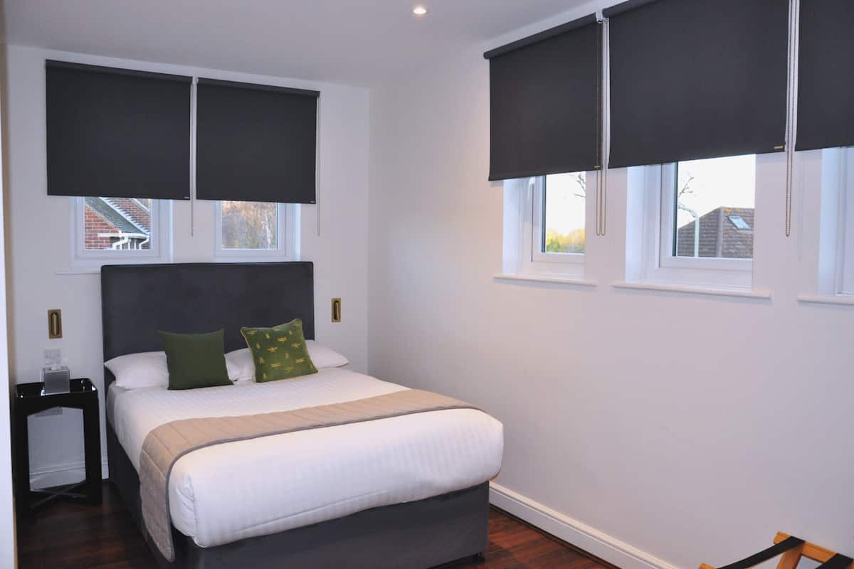 A bedroom with a bed, two windows with blinds, a nightstand, and a chair.