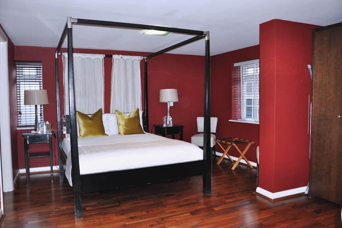 A four-poster bed with white linens and gold pillows, a bedside table with a lamp, a small folding chair, and a wooden floor.