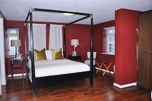 A four-poster bed with white linens and gold pillows, a bedside table with a lamp, a small folding chair, and a wooden floor.