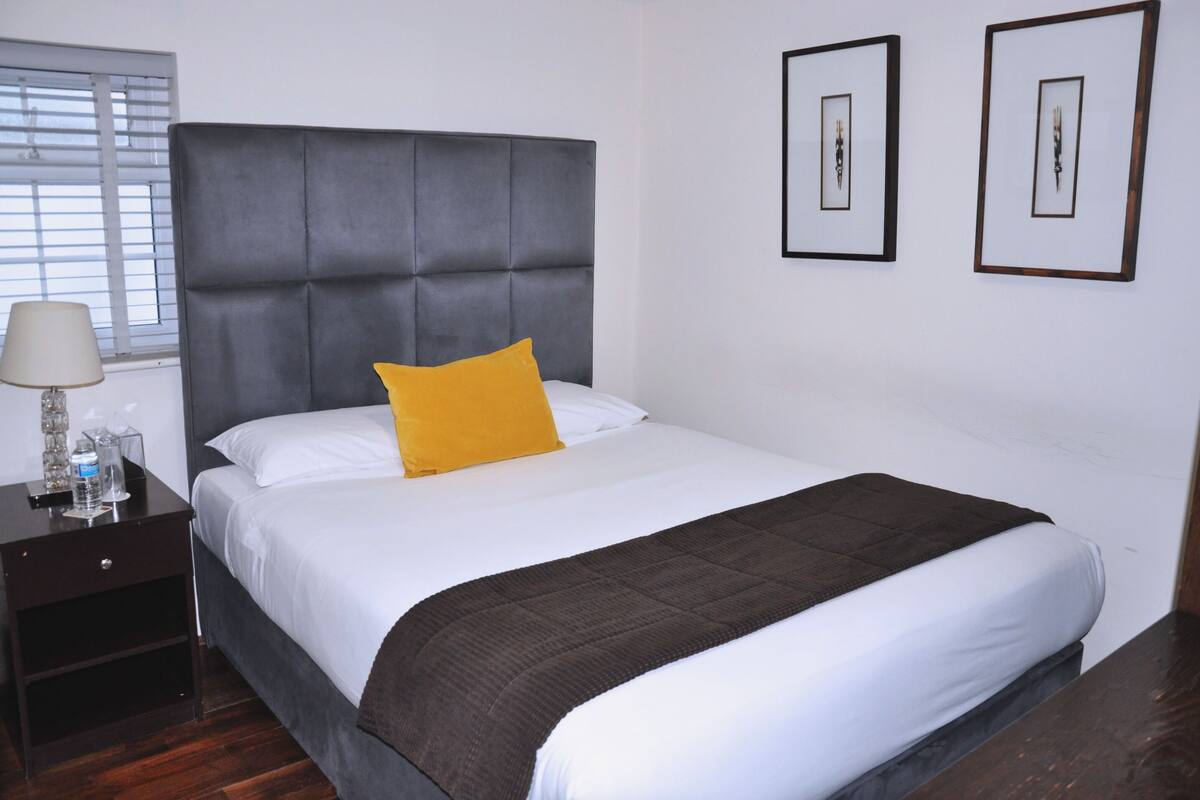 A neatly made bed with a grey headboard, a yellow pillow, and a brown throw blanket. There are two framed pictures on the wall, a nightstand with a lamp, and a window with blinds.