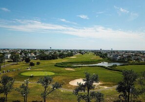 Ein Golfplatz mit Sandbunkern, einem Teich und Wohnhäusern im Hintergrund.