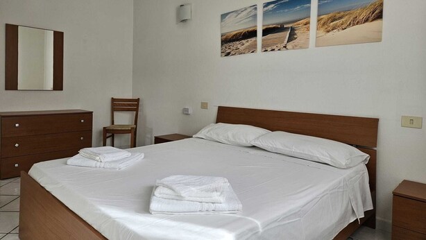 Habitación de hotel con cama, cómoda de madera, silla, mesita de noche y lámpara de pared.
