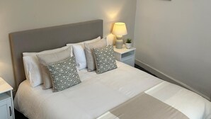 A neatly made bed with a grey headboard, white bedding, and patterned pillows. There is a bedside lamp on a nightstand, and two framed pictures on the wall. - Herring House -Only  2 minute walk  from the beach front . (Spittal)