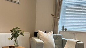 A living room with a grey sofa, a small wooden side table with a vase, and a radiator. - Herring House -Only  2 minute walk  from the beach front . (Spittal)
