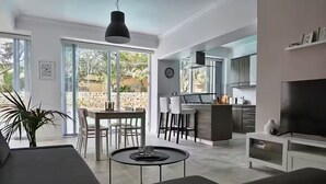 A modern living room with a sofa, a dining table with chairs, a kitchen area with a bar, and a large TV on a stand. - Villa Marni - Perfume (Munxar)