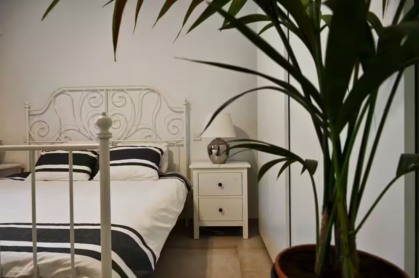 A neatly arranged bedroom with a metal-framed bed, a bedside table with a lamp, and a potted plant.