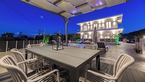 An outdoor patio with a dining table and chairs, a pool, and a modern house in the background. - Athenia Apartments (Zakynthos)