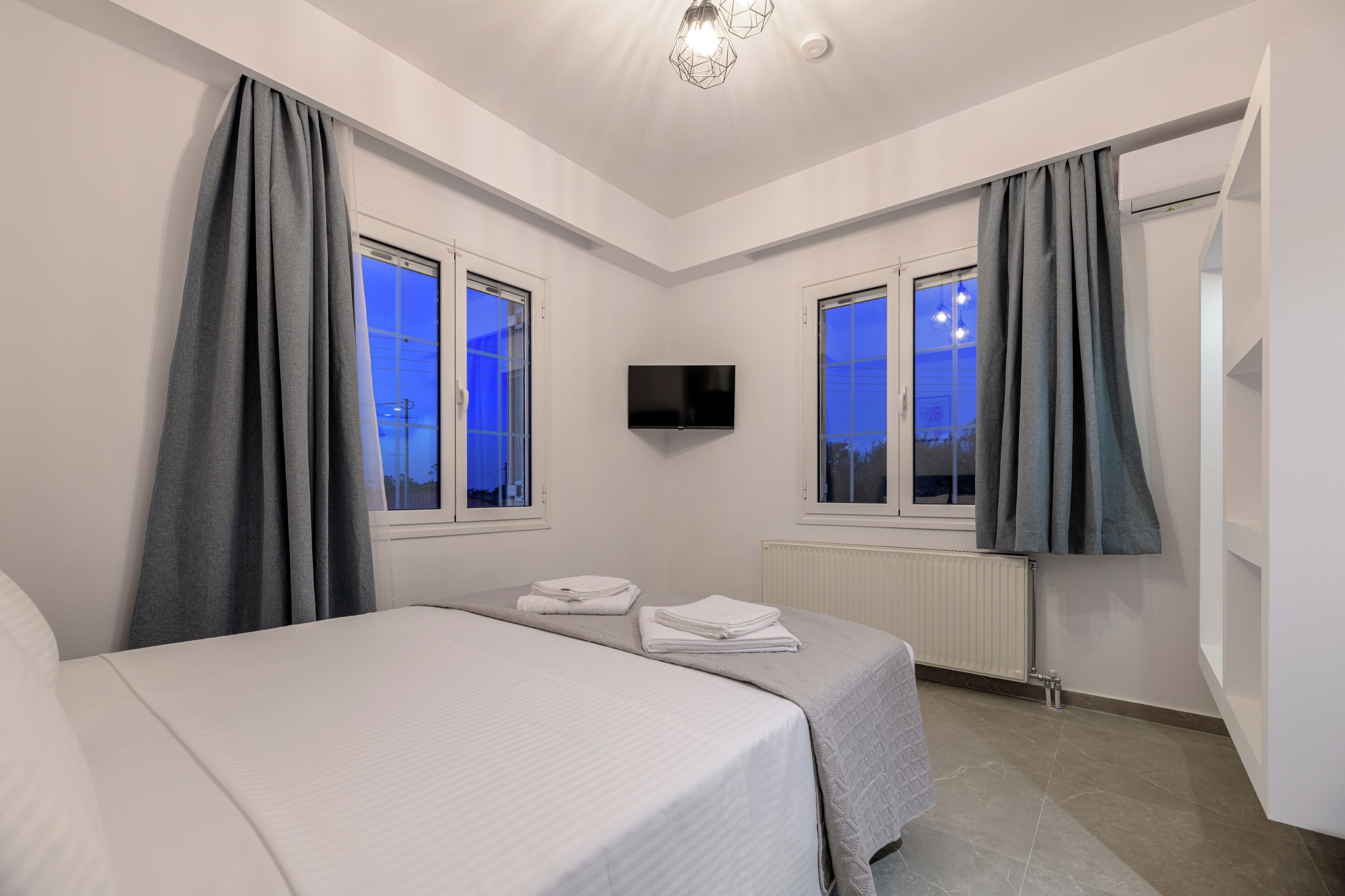 A hotel room with a bed, two windows, a flat-screen TV, and a radiator.