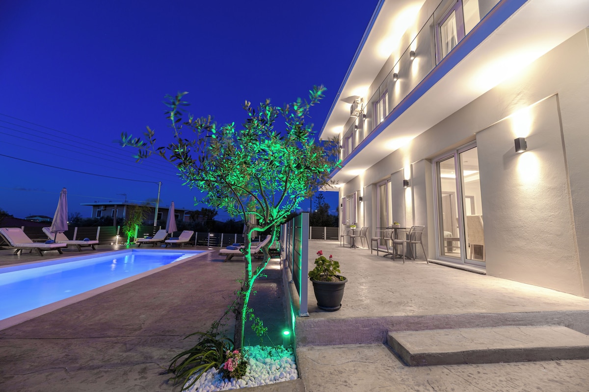 A swimming pool with lounge chairs, a potted plant, and a building with a lighted exterior.
