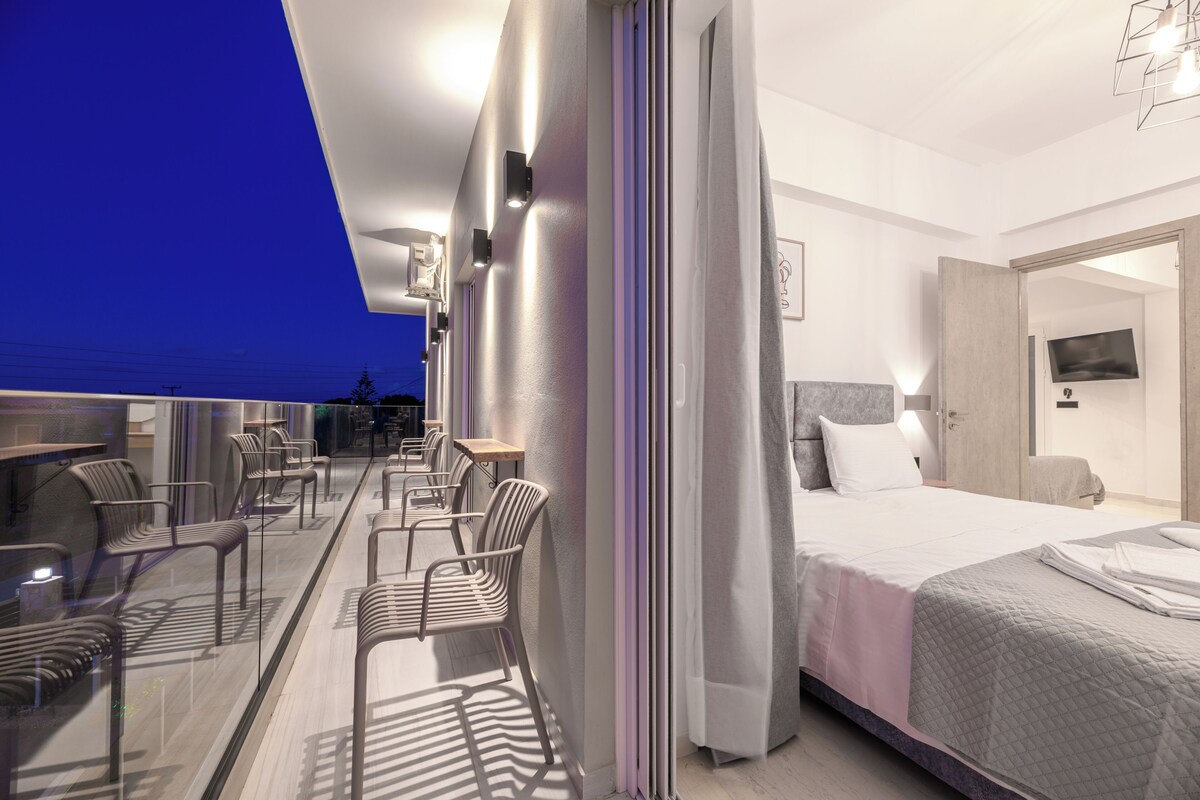 A modern hotel room with a balcony, a bed, a desk, and chairs.