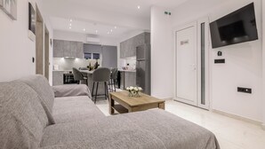 A modern hotel room with a sofa, a wooden coffee table, a TV mounted on the wall, and a kitchen area in the background. - Athenia Apartments (Zakynthos)