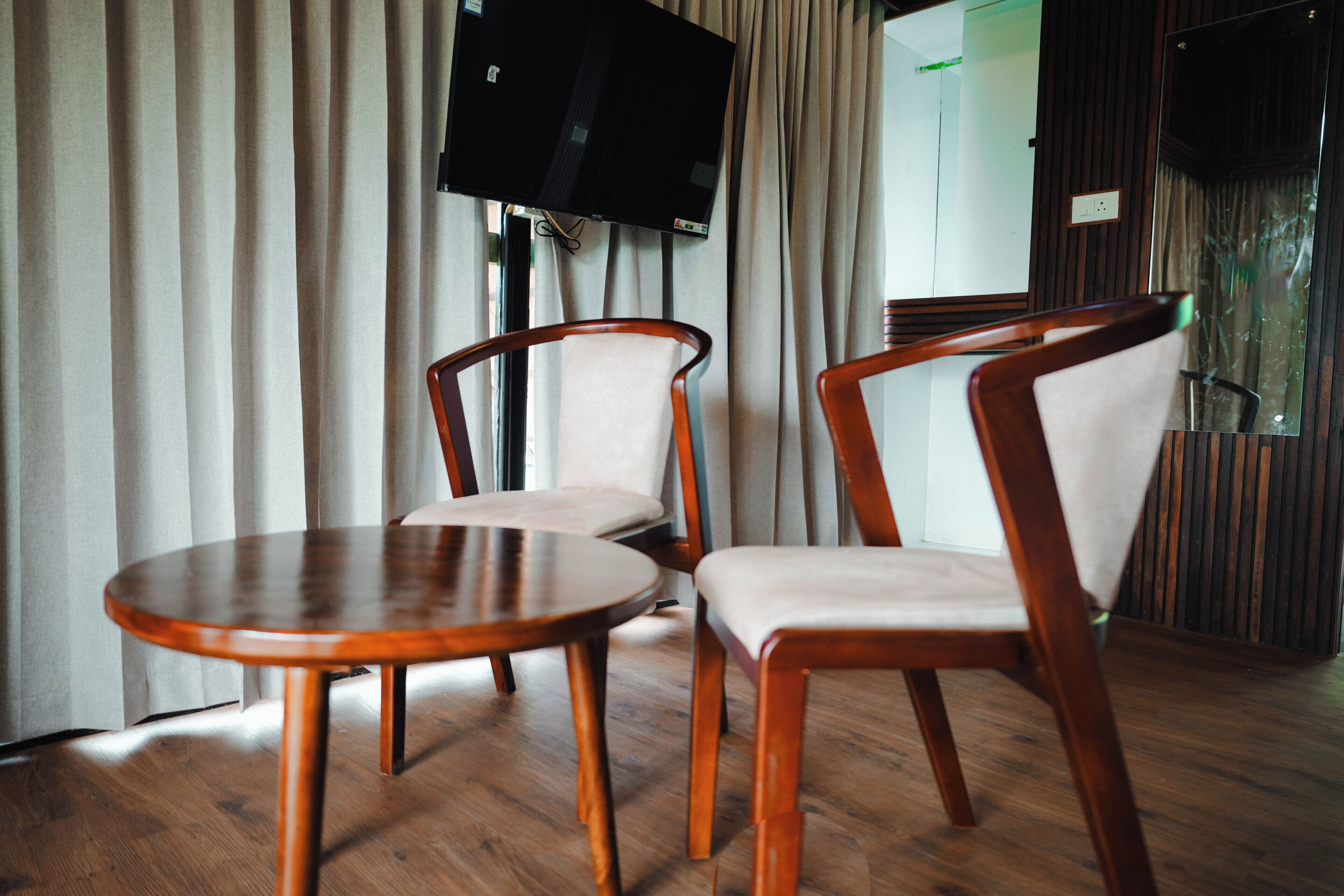 A room with two wooden chairs, a small round table, a television mounted on the wall, and a mirror reflecting part of the room.