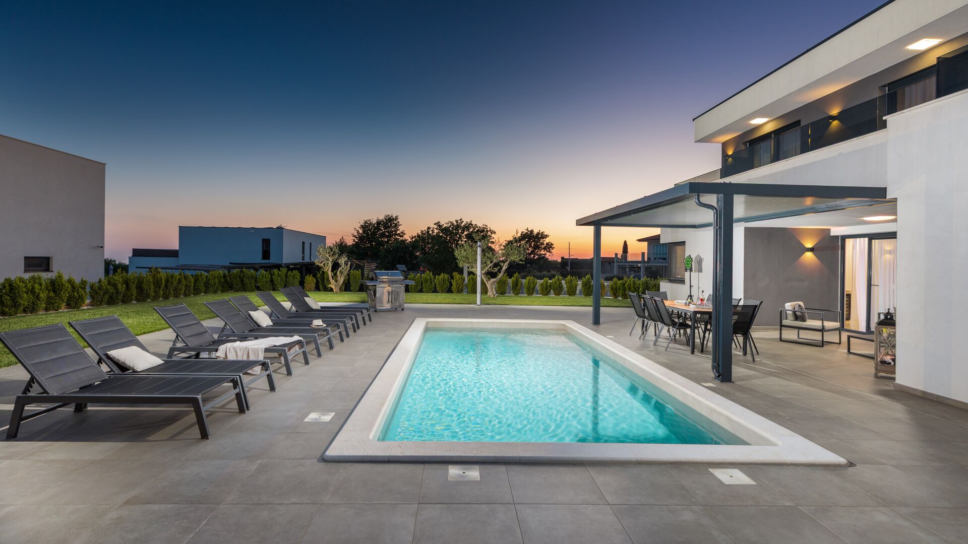 Ein moderner Poolbereich im Freien mit Liegestühlen, einem überdachten Sitzbereich und freiem Blick auf den Sonnenuntergang.