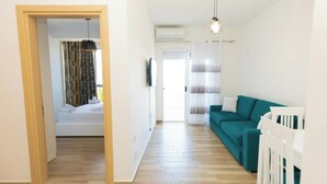 A hotel room with a wooden floor, a teal sofa, a white crib, and a bed with a curtain. - Seaview Apartments (Orikum)