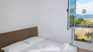A neatly made bed with white linens and towels, a brown headboard, and a large window with a view of a landscape. - Seaview Apartments (Orikum)