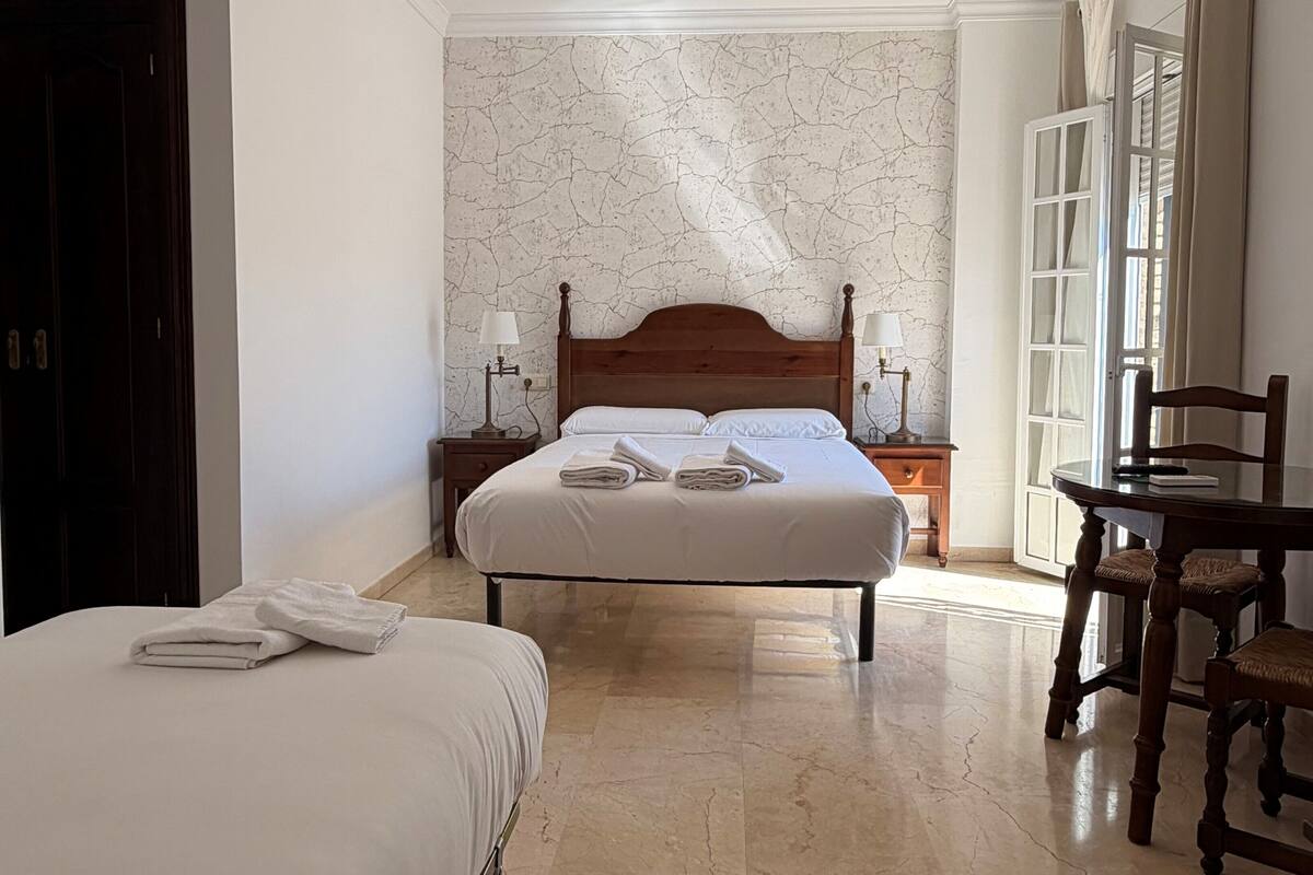 Habitación de hotel con una cama grande, una cama más pequeña, una silla de madera, una mesa y una mesita de noche con lámparas.
