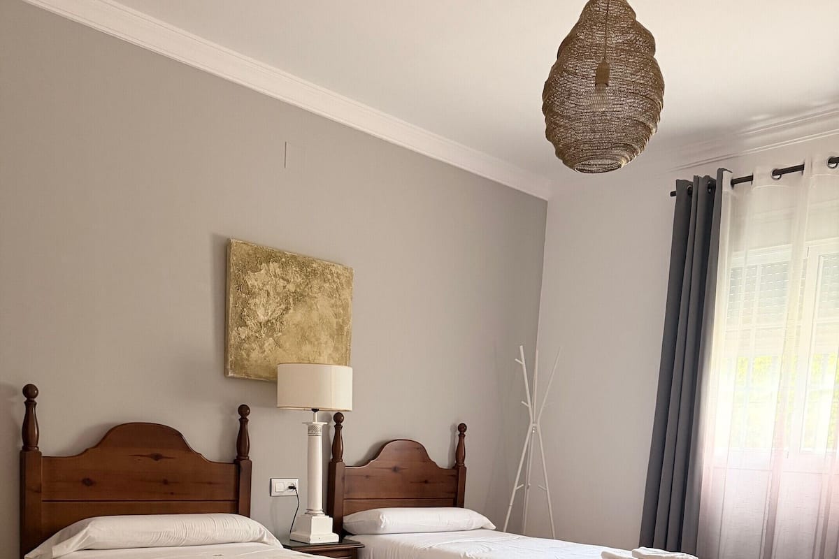 Una habitación con dos camas de madera, una lámpara de pie, un cuadro en la pared y una ventana con cortinas.