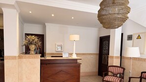 A reception area with a wooden reception desk, a framed certificate, a lamp, a vase with flowers, and a warning sign on the wall. - Hotel Victoria Matalascañas (Matalascañas)