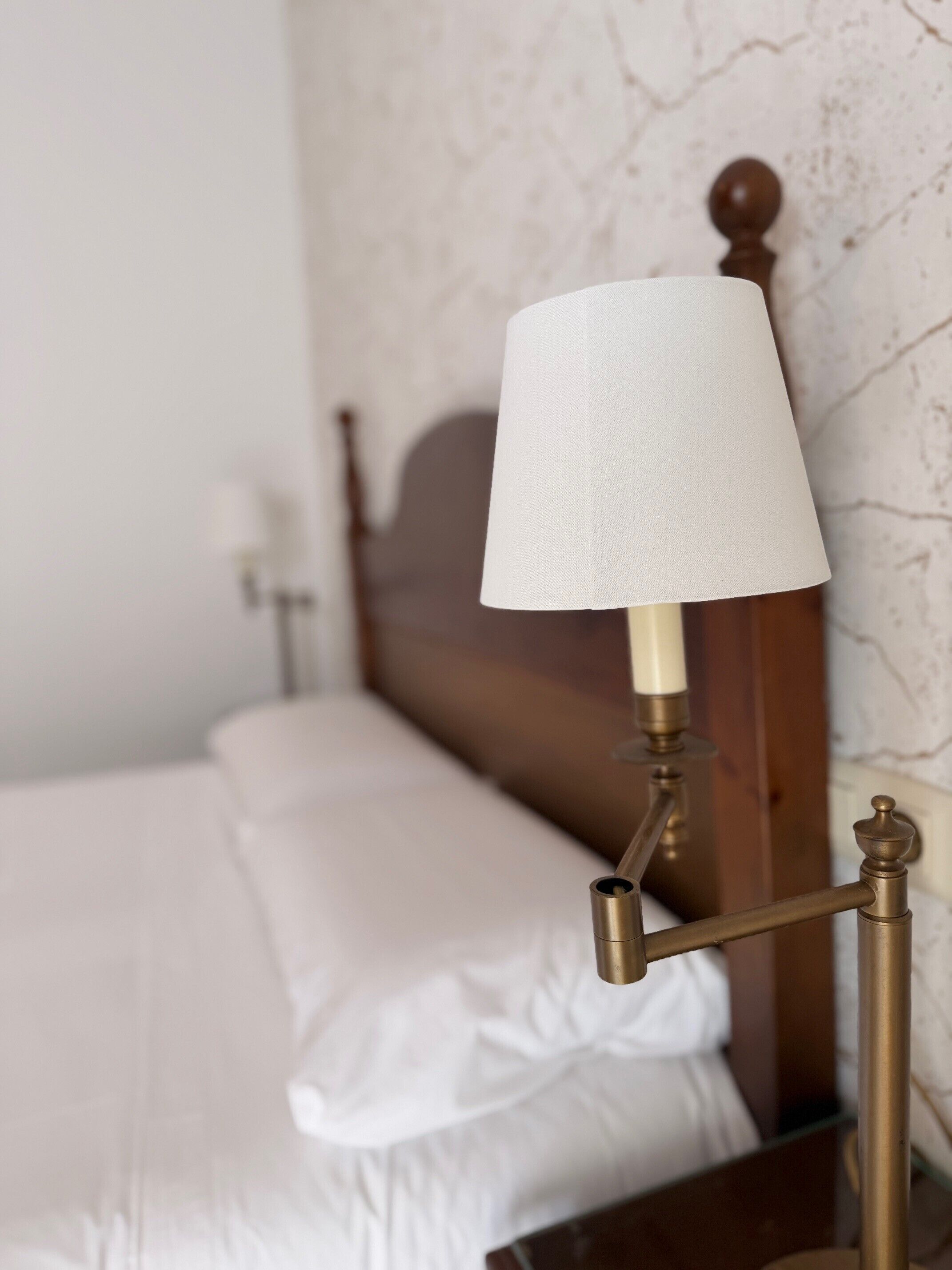 Ein Bett mit weißer Bettwäsche, ein Holz-Kopfteil und ein Messing-Nachttisch mit weißem Lampenschirm.