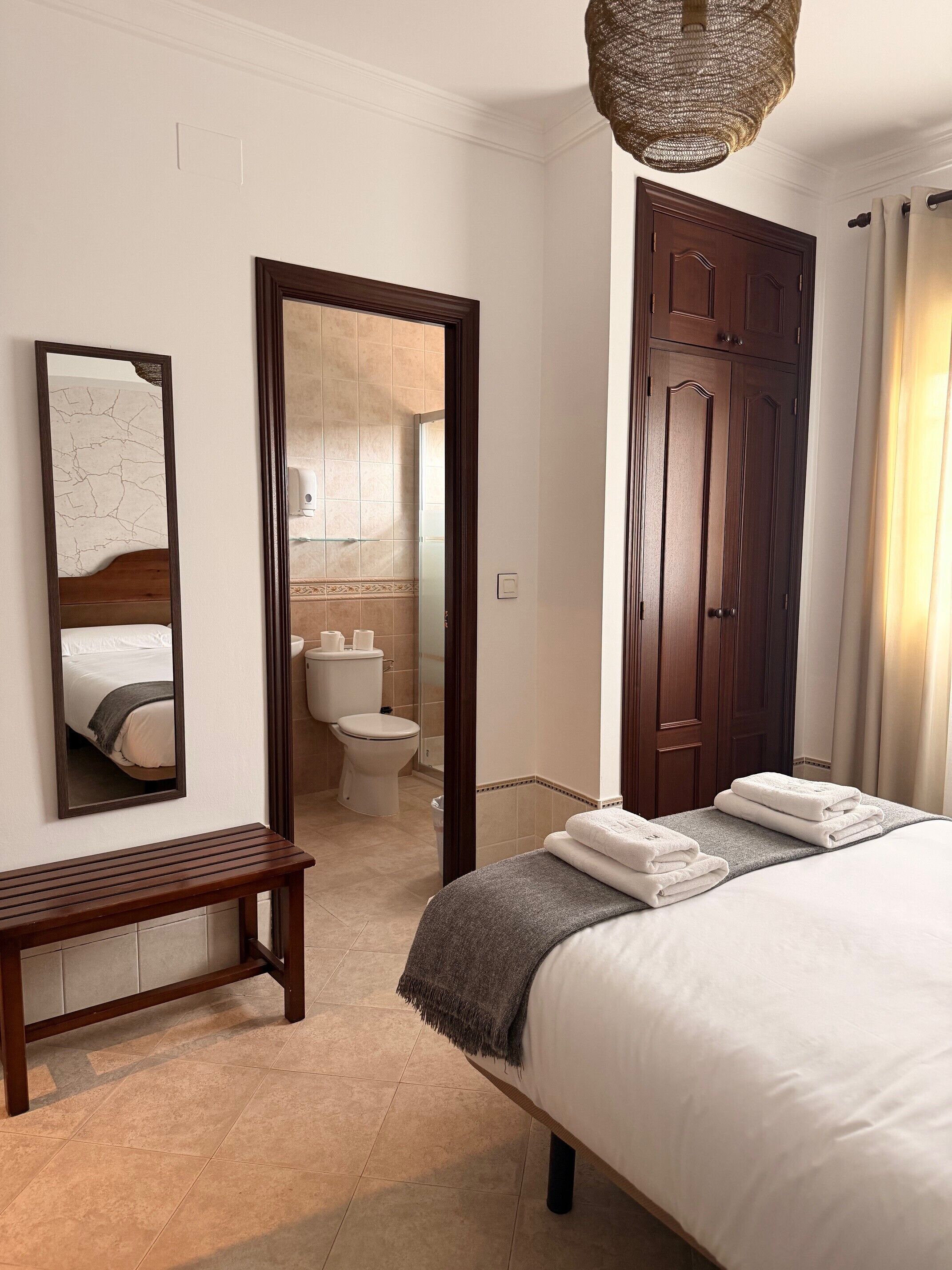 Ein Hotelzimmer mit Bett, einer Holzbank, einem Badezimmer mit Toilette und Waschbecken sowie einem großen Spiegel.