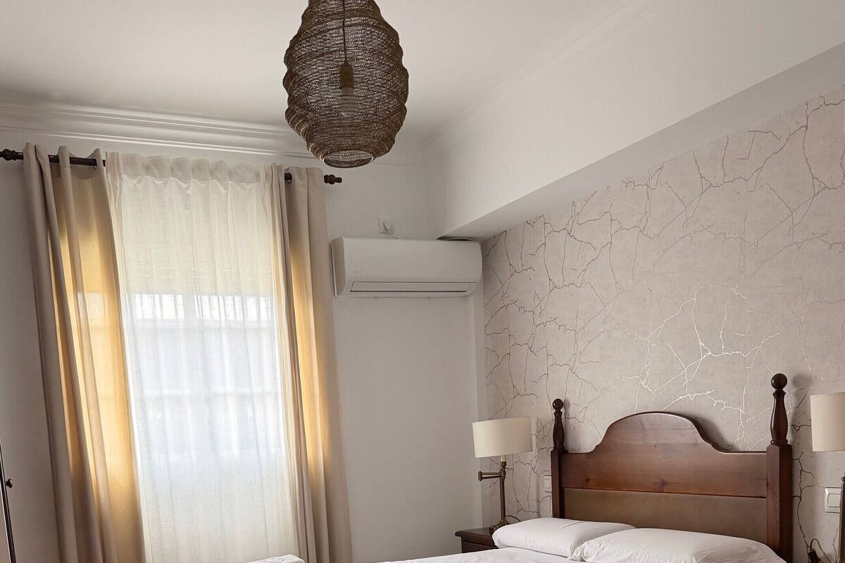 Un dormitorio con cama, mesitas de noche, lámpara, lámpara colgante y ventana con cortinas.