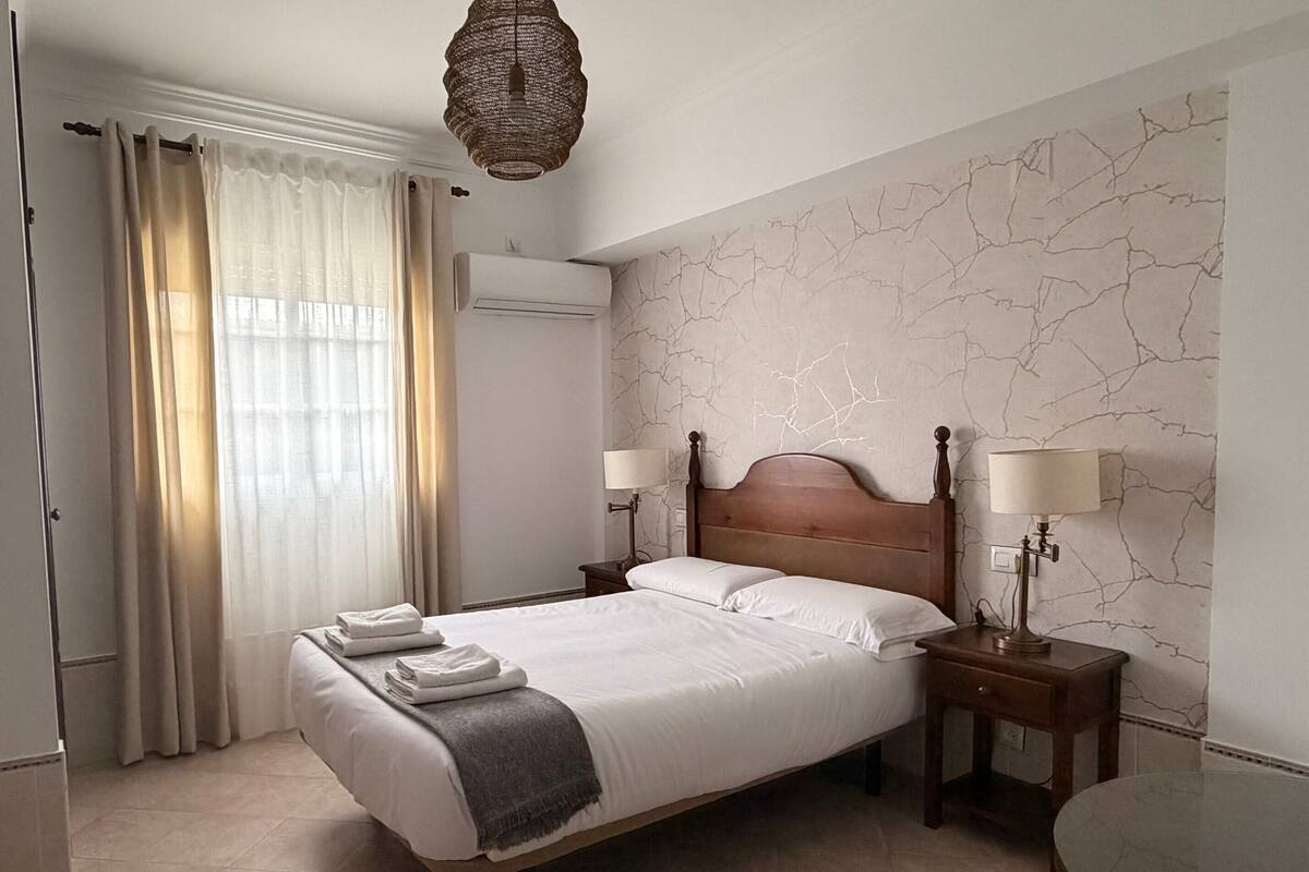 Una habitación de hotel con cama, dos mesitas de noche con lámparas, una ventana con cortinas, un equipo de aire acondicionado y una pared con diseño de mármol.