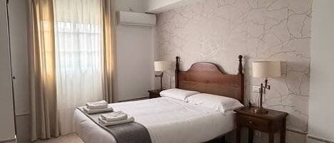 Een hotelkamer met een bed, twee nachtkastjes met lampen, een raam met gordijnen, een airconditioning en een wand met marmer dessin.