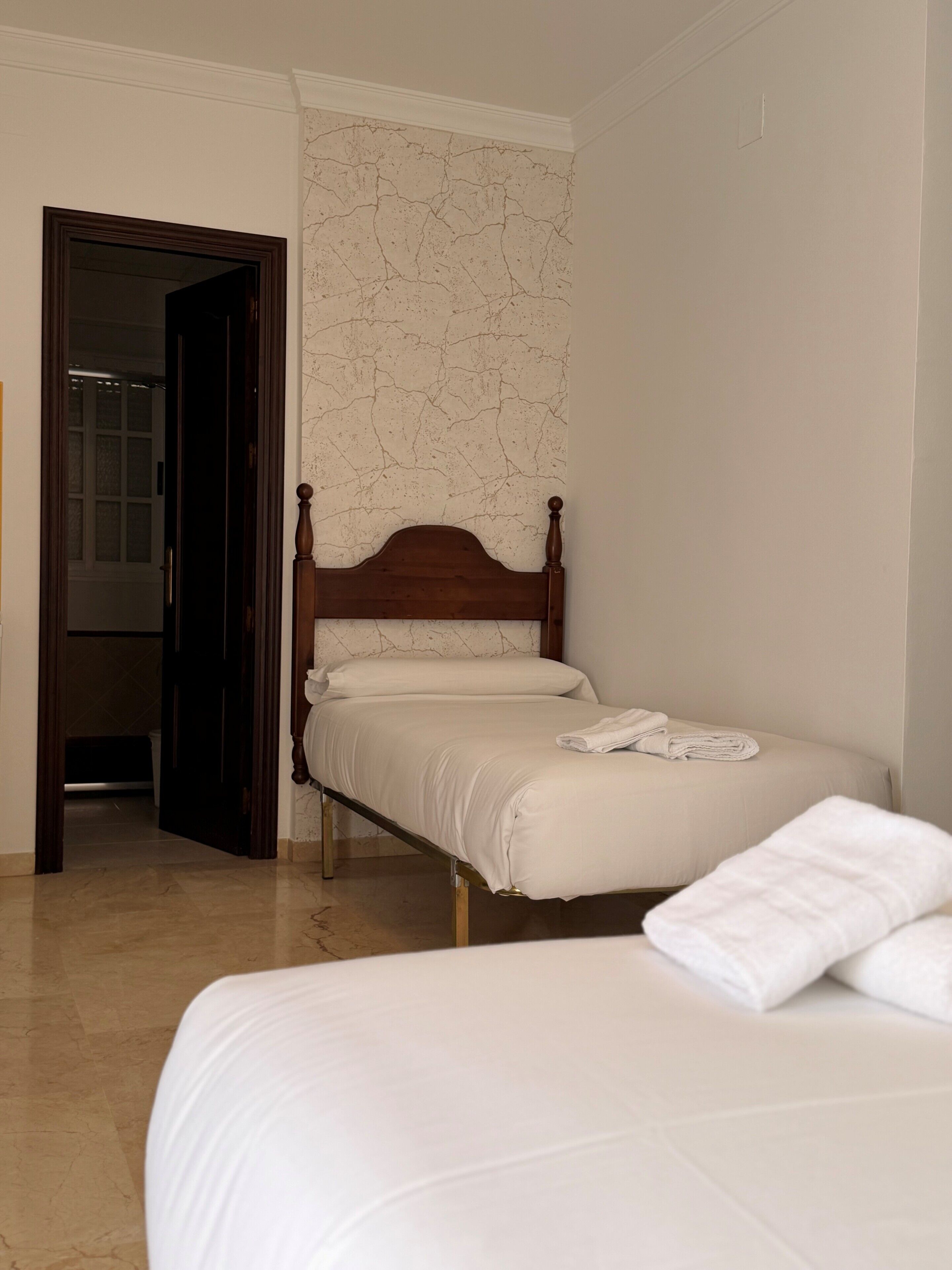 Quarto com cama de madeira, parede branca com textura rachada e porta que dá para outro cômodo.