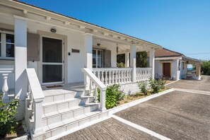 A two-story house with a covered porch, a front door, and a small garden area. - Serenity Apartments (Corfu)