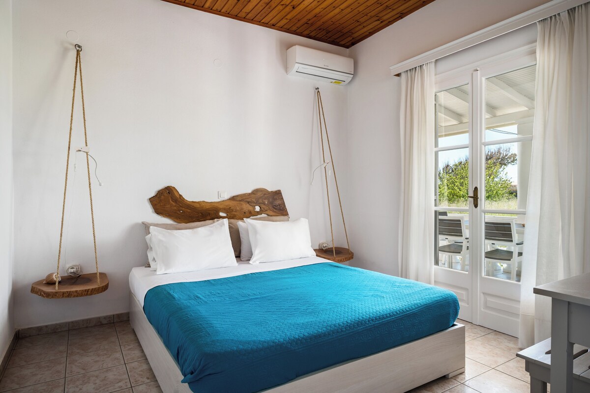 Ein Schlafzimmer mit Bett, weißen Wänden, einem hölzernen Kopfteil, einer Hängeschaukel und Blick ins Freie durch eine Glastür.